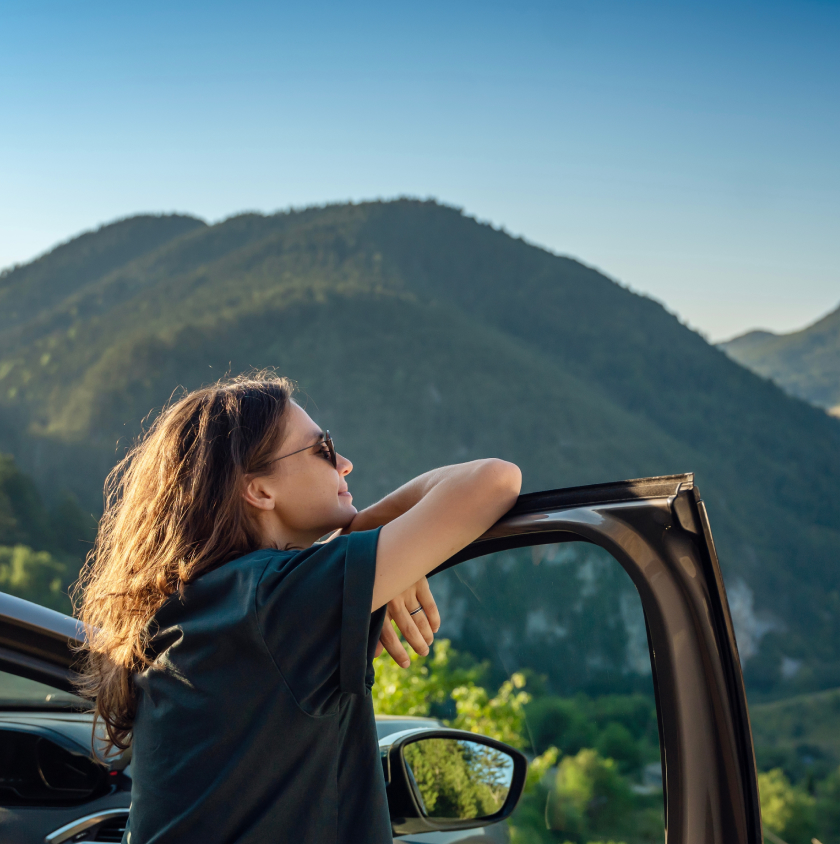 Relaxed-young-woman-enjoying-scenic-mountain-view-from-car-door-wearing-sunglasses-on-a-sunny-day